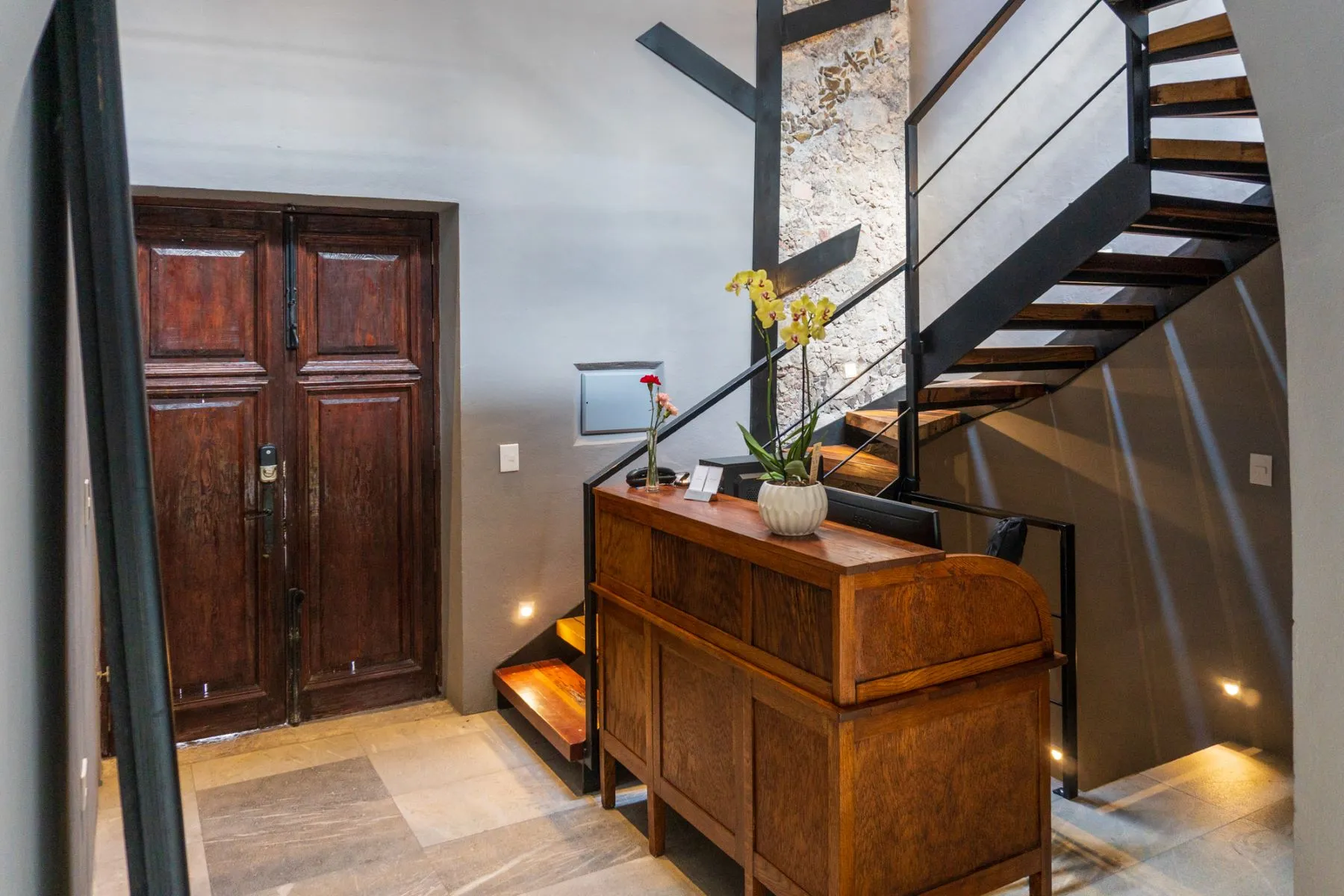 Floating steel and wood staircase against an exposed stone wall beside an antique rolltop desk